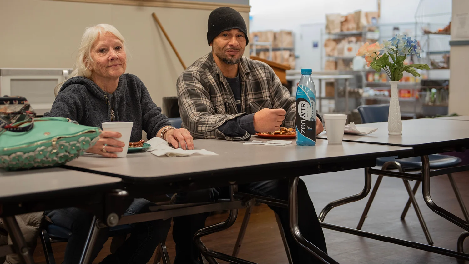 At a Catholic Charities housing site, a man with a beard in a fleece vest bends over a washing machine, checking on his clothes. Another man, wearing a purple hoodie, sits nearby.